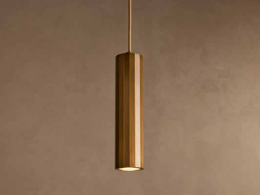 A cylindrical pendant light with a glossy brass finish hangs from the ceiling against a plain beige background.