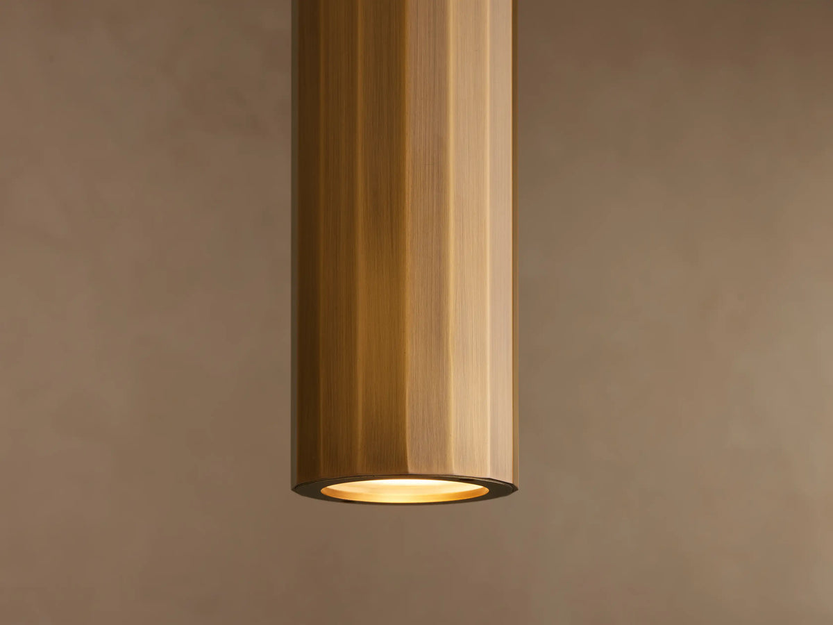 Sleek cylindrical brass pendant light hanging against a textured beige wall, featuring a polished reflective surface.