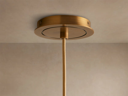 A cylindrical brass pendant light with a smooth, brushed finish, hanging downward against a neutral beige background, illuminated warmly.