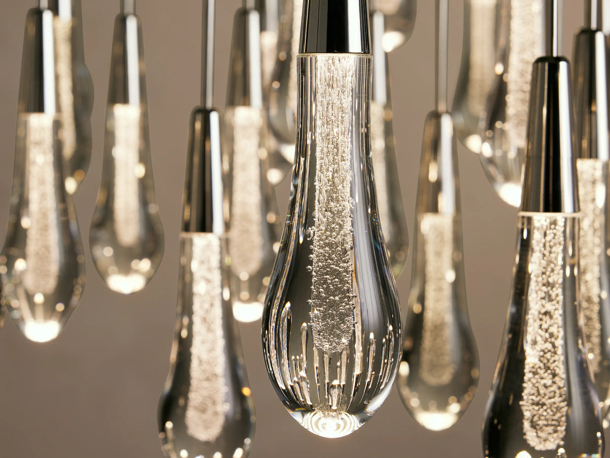 Teardrop-shaped glass pendants hang from a Soltaire Round Chandelier, each with a silver metallic top, emitting a warm light.
