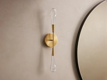 Soltaire Double Sconce with a brass finish, two elongated LED bulbs vertically aligned on a circular backplate against a beige wall.