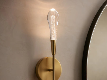 Soltaire Double Sconce with brass finish, featuring two clear elongated bulbs, mounted on a round wall plate against a neutral wall.