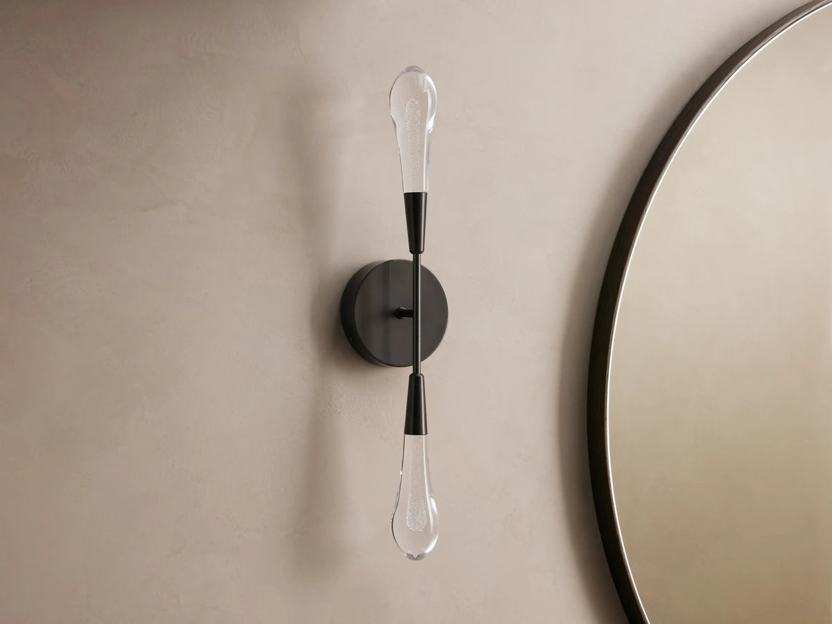Soltaire Double Sconce with a black steel backplate and elongated teardrop glass bulbs, mounted on a beige wall near a round mirror.