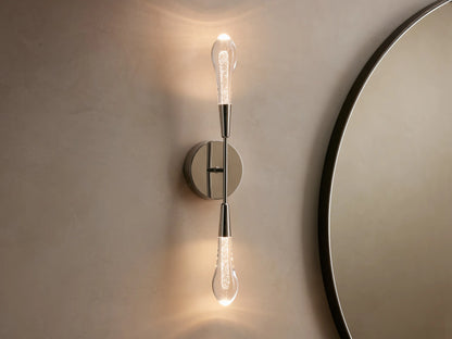 Soltaire Double Sconce mounted on a wall, features two teardrop LED bulbs on a polished nickel backplate, emitting warm light.