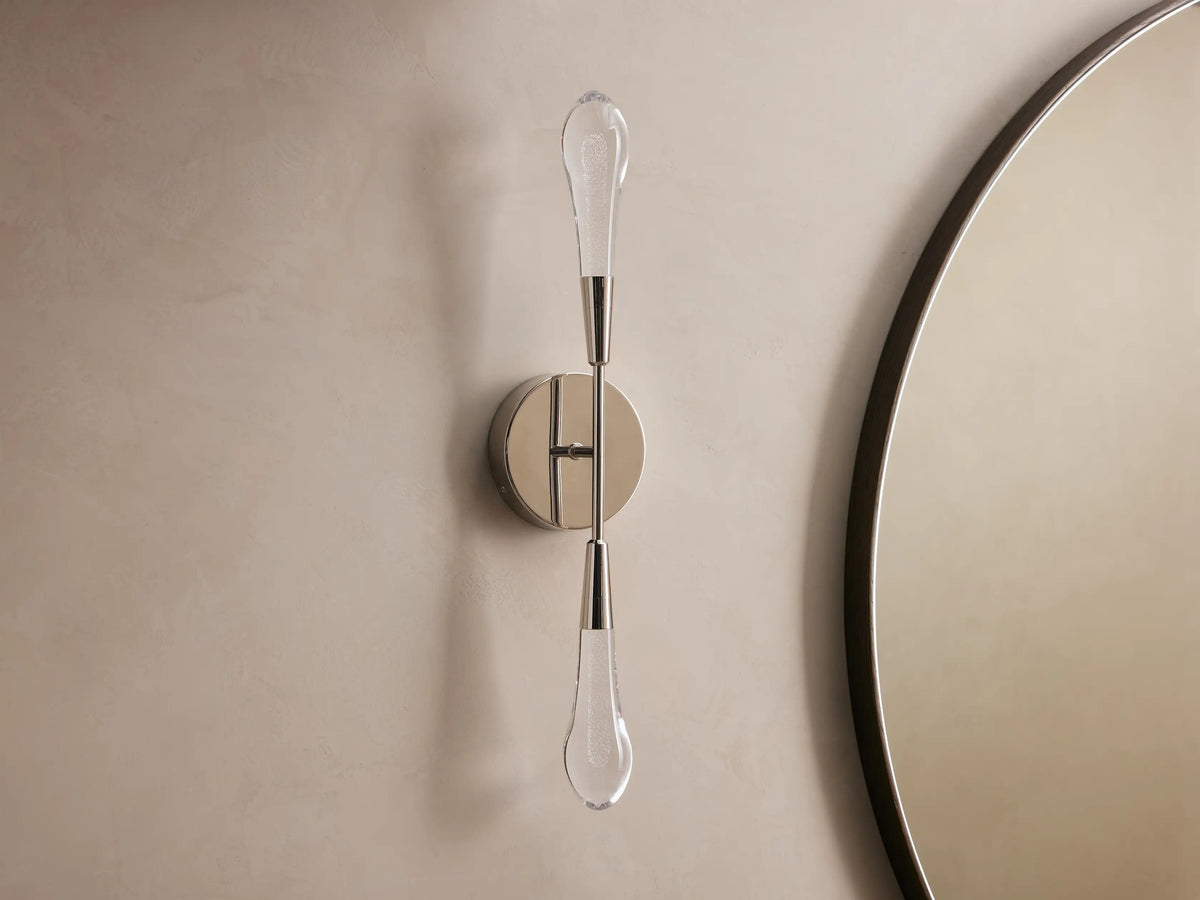 Soltaire Double Sconce mounted on a wall, features two teardrop LED bulbs on a polished nickel backplate, emitting warm light.