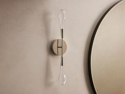 Soltaire Double Sconce mounted on a wall, features two teardrop LED bulbs on a polished nickel backplate, emitting warm light.