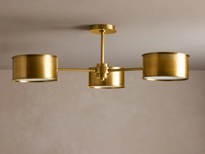 Daphne Metal Flush Mount with three antique brass cylindrical shades, glowing warmly, mounted on a light ceiling.