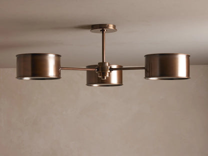 Daphne Metal Flush Mount in warm bronze with three cylindrical shades casting a soft, warm glow on a light ceiling.