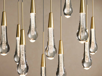 Soltaire Linear Chandelier with multiple glowing, teardrop-shaped clear glass pendants filled with bubbles, suspended at varying heights from brass rods.