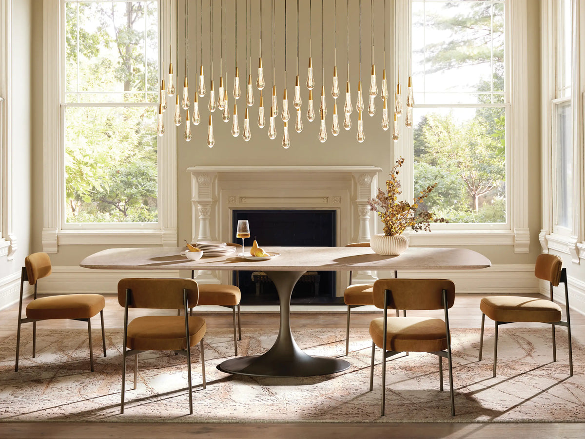 Soltaire Linear Chandelier with many illuminated, clear teardrop-shaped pendants featuring sparkling seeded glass, against a warm brown background.