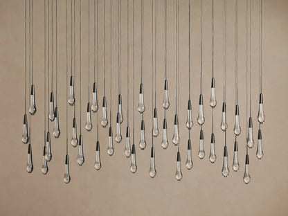 Soltaire Linear Chandelier features multiple clear glass teardrop pendants with dark tops, suspended at varied heights on beige.