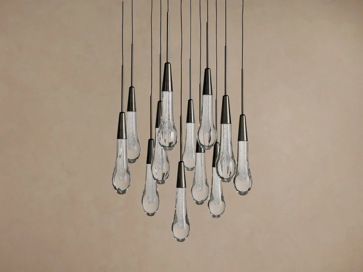 Soltaire Round Chandelier with multiple clear, textured teardrop glass pendants and dark metal caps, hanging against a beige wall.