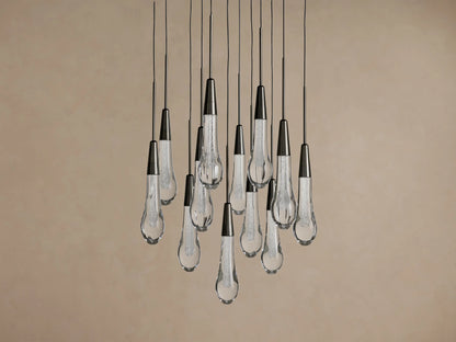 Soltaire Round Chandelier with multiple clear, textured teardrop glass pendants and dark metal caps, hanging against a beige wall.