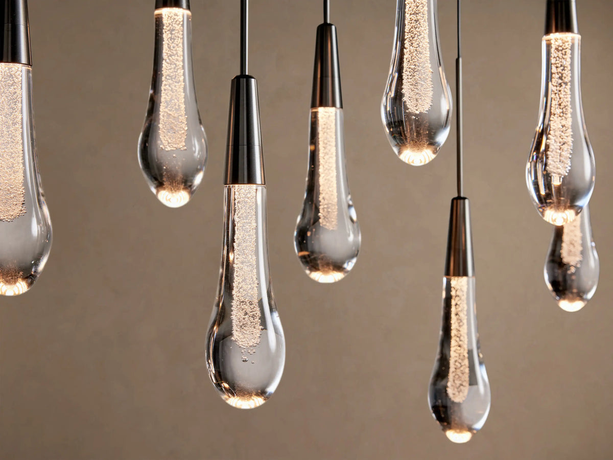 Soltaire Linear Chandelier: illuminated clear glass teardrop pendants with sparkling internal bubbles & dark caps, hanging.