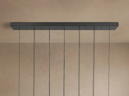 Soltaire Linear Chandelier showing a dark gray linear ceiling mount with multiple black cords hanging down against a light brown wall.