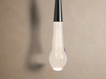 Soltaire Sconce: Dark bronze circular backplate on a tan wall, with a slender rod holding a clear bubbled glass teardrop.
