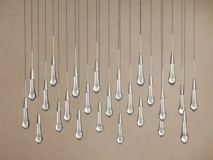 Clear Soltaire Linear Chandelier with numerous illuminated teardrop glass pendants, silver caps, and dark cords against a brown background.
