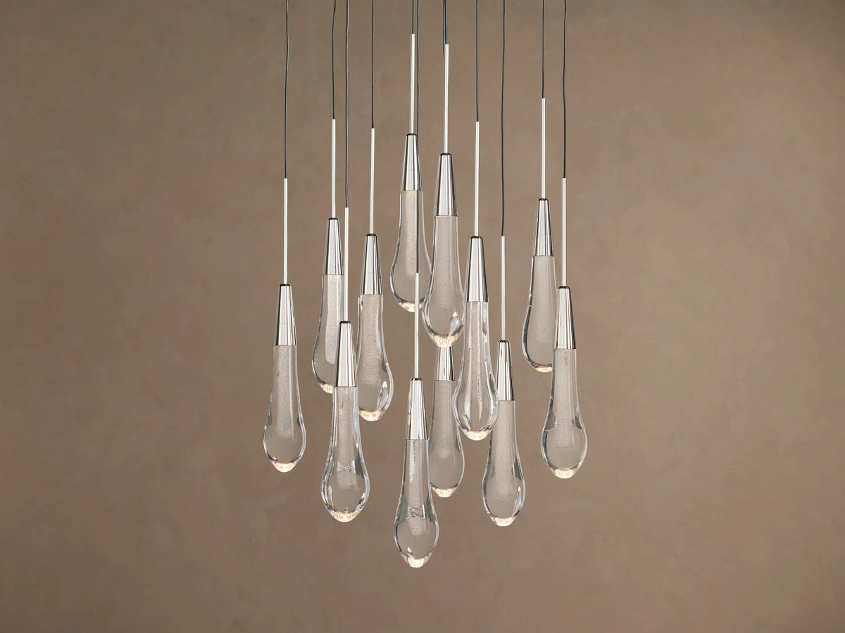 Soltaire Round Chandelier: Clear glass teardrop pendants with metallic caps, glowing, hanging at varied heights on brown background.