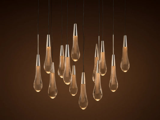 Soltaire Round Chandelier: Clear glass teardrop pendants with metallic caps, glowing, hanging at varied heights on brown background.