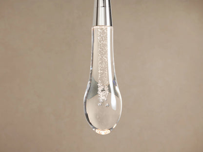 Soltaire Pendant: clear glass teardrop fixture with internal bubbles, silver top & stem, hanging vertically against a beige wall.