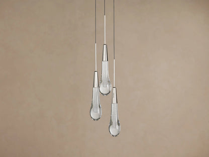 Three Soltaire Cluster Pendants with clear tear-drop glass shades, silver caps, and glowing textured interiors hanging against a brown wall.