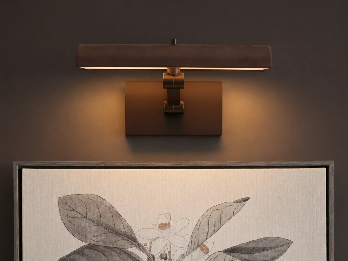 Abel Picture Light Sconce, a dark bronze curved light bar with warm LED light, mounted on a wall above a framed botanical artwork.