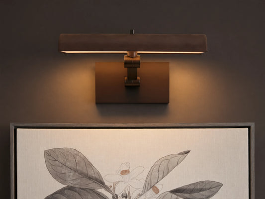 Abel Picture Light Sconce, a dark bronze curved light bar with warm LED light, mounted on a wall above a framed botanical artwork.