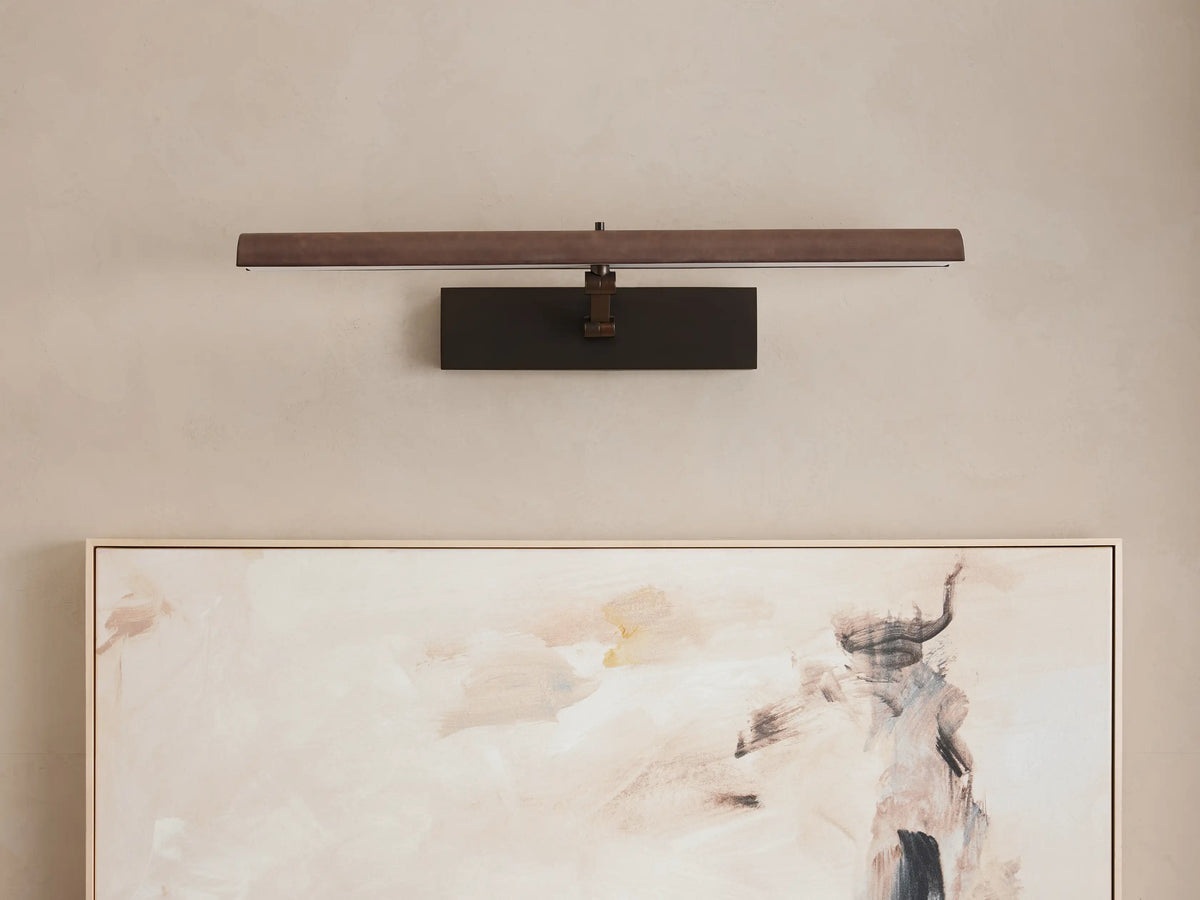 Abel Picture Light Sconce with a dark bronze finish, mounted horizontally on a light wall, casting a warm glow onto an abstract painting.