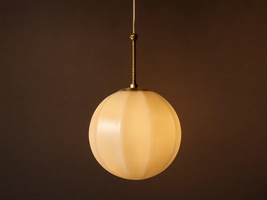 Agnes Pendant: Spherical cream paneled glass shade illuminated, hanging from ribbed antique brass rod against a gray wall.
