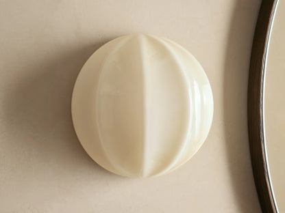 Agnes Clamshell Sconce: Spherical cream paneled glass sconce, glowing warmly, mounted on a light wall next to a dark mirror.