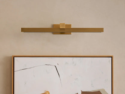 Corinth Picture Light Sconce, brushed brass, rectangular bar design with illuminated LED strip, mounted horizontally above abstract art.