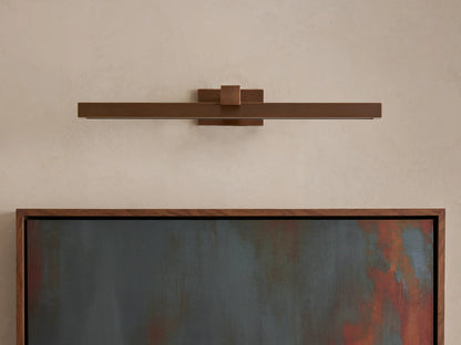 Corinth Picture Light Sconce, a long, horizontal bronze fixture on a light wall, casting warm LED light onto a framed abstract painting.