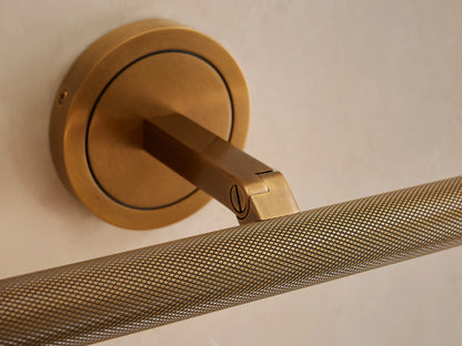 Close-up of an antique brass Monroe Picture Light Sconce mounted on a light wall, featuring a knurled textured light bar.