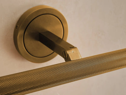 Antique brass Monroe Picture Light Sconce with round base, arm, and horizontal knurled light bar, against a cream wall.
