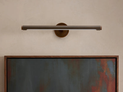 Monroe Picture Light Sconce, horizontal, knurled antique brass, lighted, above a framed abstract painting.