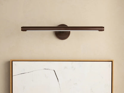 Monroe Picture Light Sconce in antique brass, mounted on a beige wall, illuminating an abstract artwork with a wood frame.