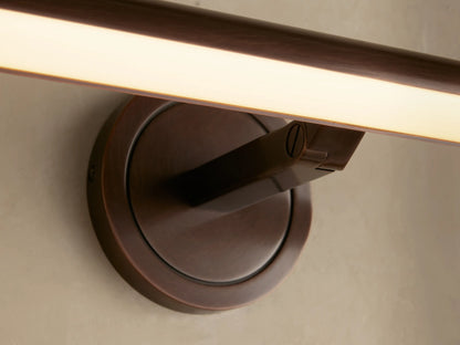 Close-up of Monroe Picture Light Sconce in antique brass. The arm extends from a circular base, emitting warm LED light on a beige wall.