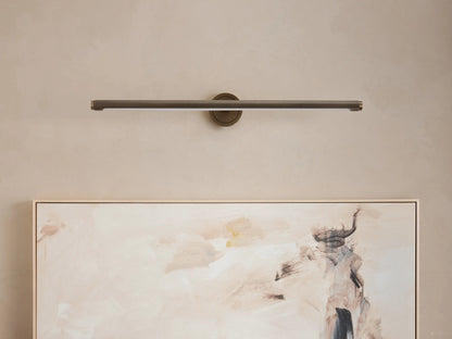 Monroe Picture Light Sconce in antique brass, a horizontal bar-style light mounted on a beige wall above an abstract painting.