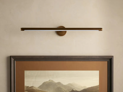 Antique brass Monroe Picture Light Sconce with a soft LED glow over a framed mountain landscape on a beige wall.