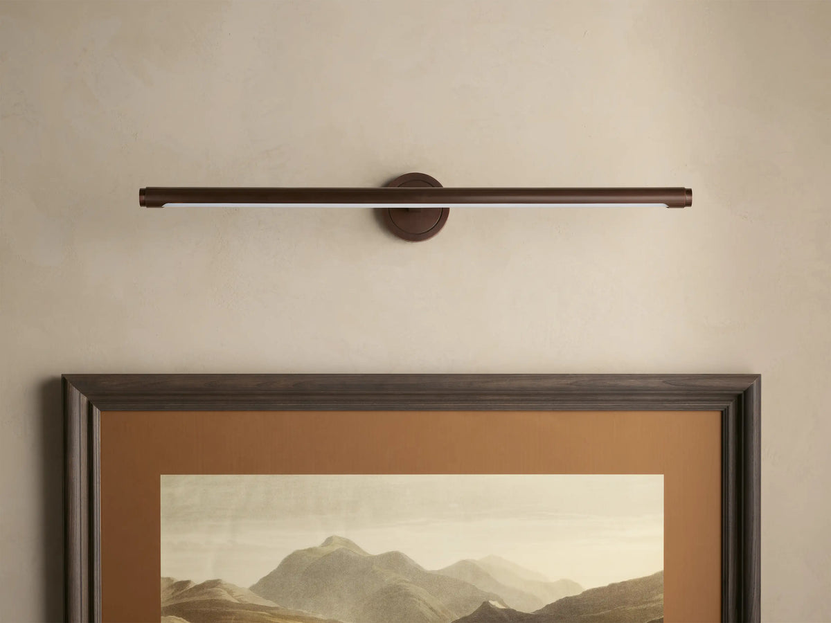 Dark bronze Monroe Picture Light Sconce, a linear bar emitting warm LED light, mounted above a framed mountain art on a beige wall.