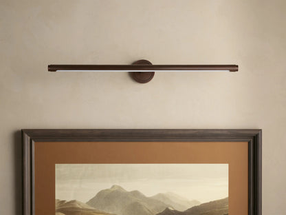 Dark bronze Monroe Picture Light Sconce, a linear bar emitting warm LED light, mounted above a framed mountain art on a beige wall.