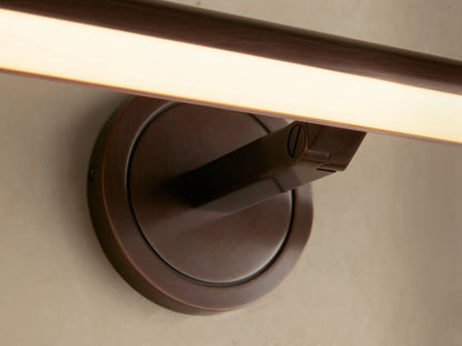 Antique brass Monroe Picture Light Sconce, mounted on a light wall, with its acrylic diffuser glowing with warm LED light.