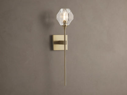 Amoroso Sconce: brass-plated wall light with long rod and illuminated faceted clear crystal shade on a brown wall.