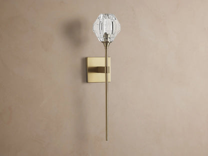 Amoroso Sconce: brass-plated wall light with long rod and illuminated faceted clear crystal shade on a brown wall.