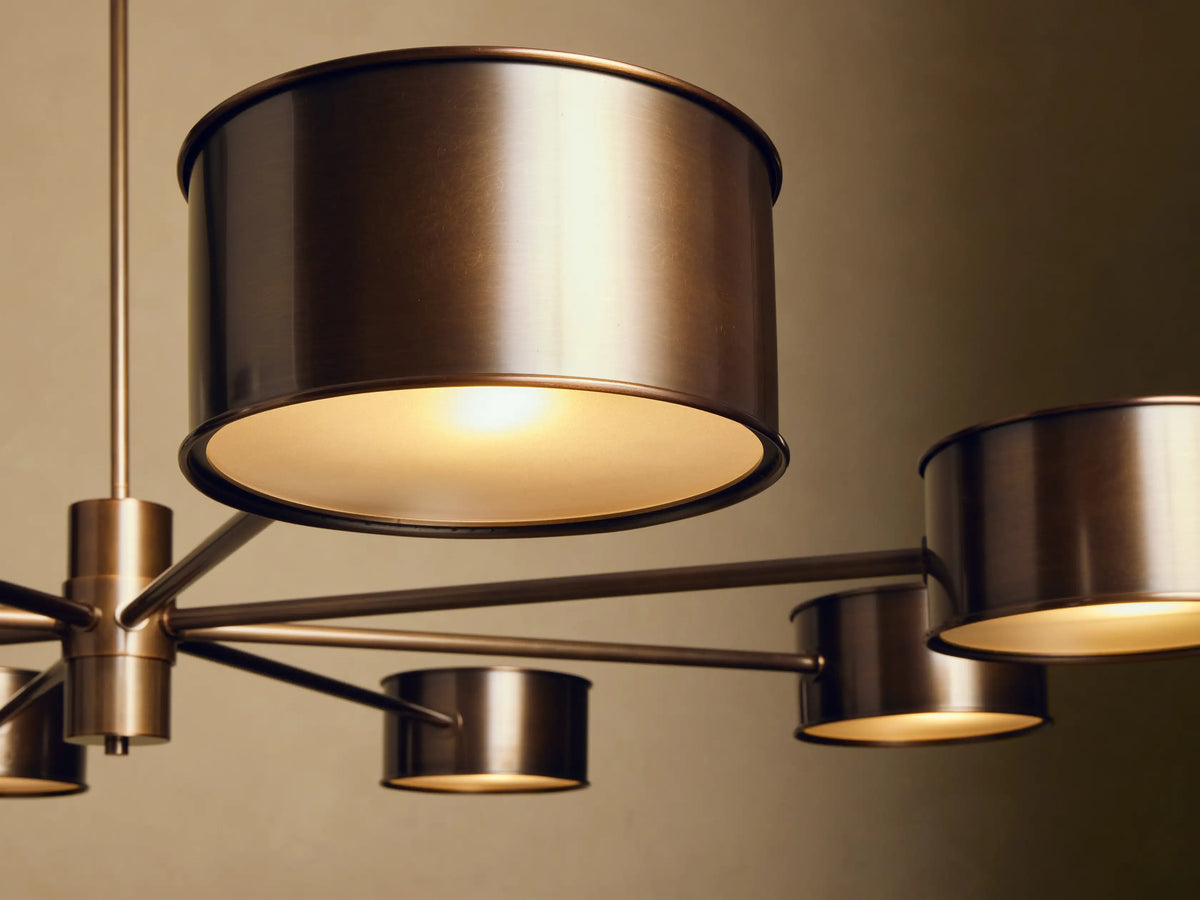 Daphne Metal Round Chandelier, warm bronze, 8 cylindrical metal shades with white diffusers, hanging against a beige wall.