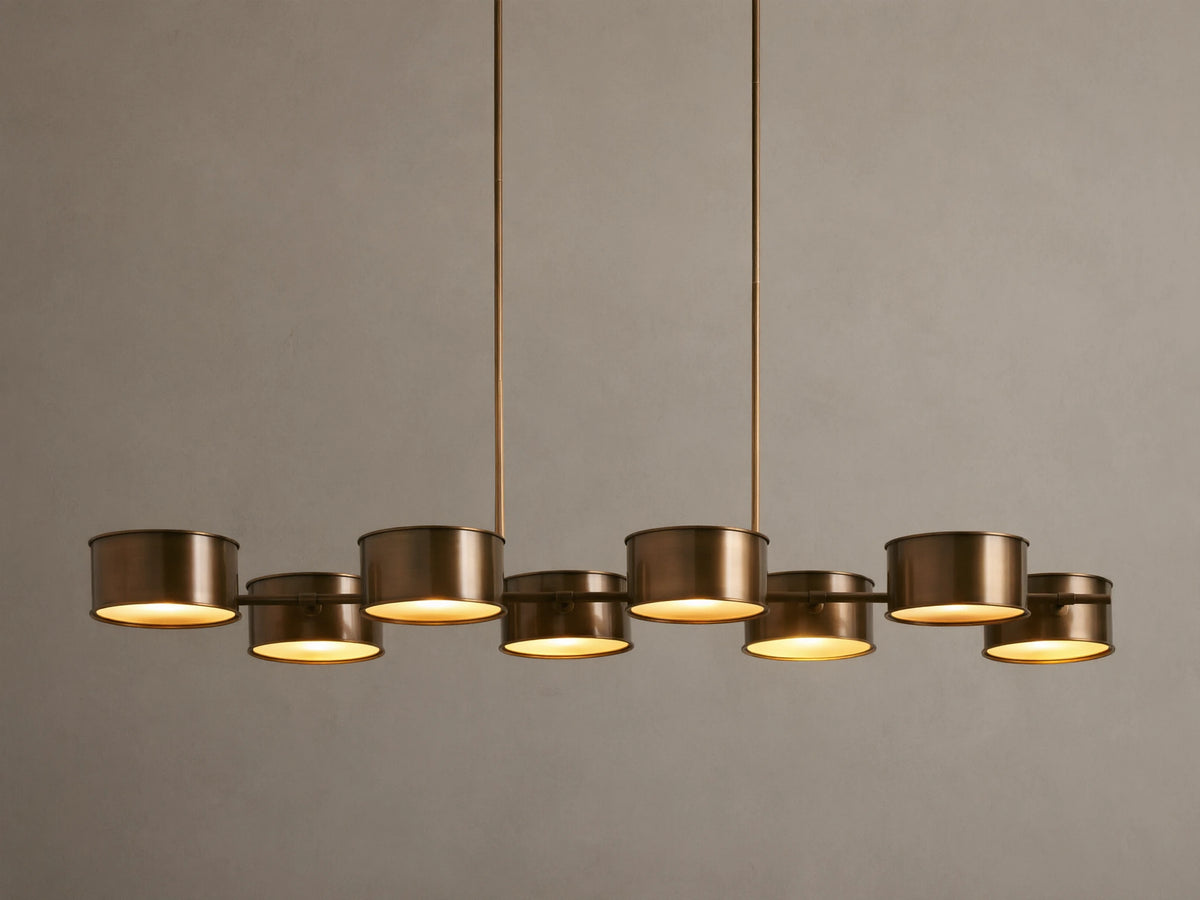 Daphne Metal Linear Chandelier with 8 warm bronze drum shades suspended from rods, emitting a soft downward glow.