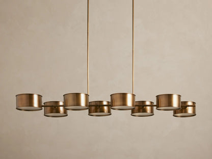 Daphne Metal Linear Chandelier with 8 warm bronze drum shades suspended from rods, emitting a soft downward glow.