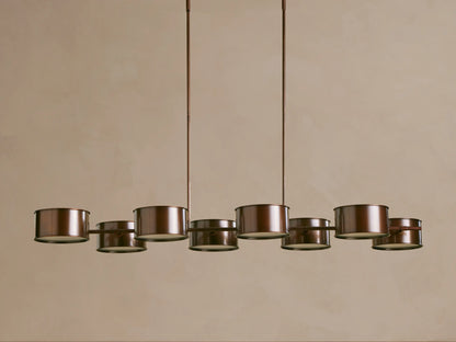 Daphne Metal Linear Chandelier, dark bronze, with 8 glowing cylindrical shades suspended from two rods against a beige wall.