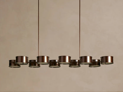 Daphne Metal Linear Chandelier with 9 dark bronze drum shades, emitting warm light, hanging from rods.