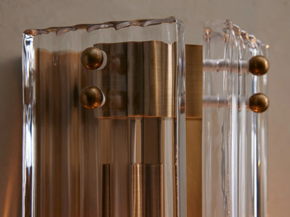 Delsie Sconce with burnished brass metalwork and fluted clear glass panels mounted vertically on a light gray wall.